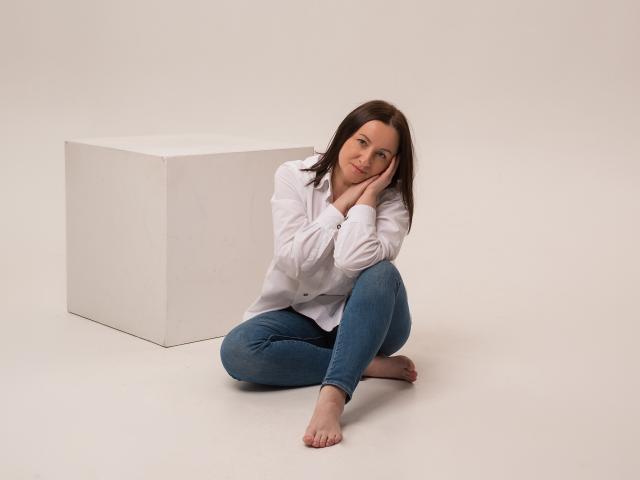 AbbyTaylorr with shoulder-length brown hair, wearing a white shirt and blue jeans, sits barefoot, leaning on her hand, next to a white cube in a studio.