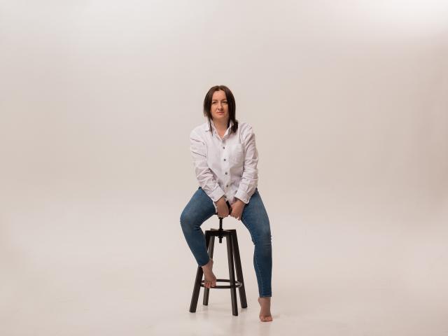 AbbyTaylorr with tits covered, sitting on a stool, wearing a white shirt, blue jeans, and bare feet, smiling slightly, hands on knees, brown hair, white background.