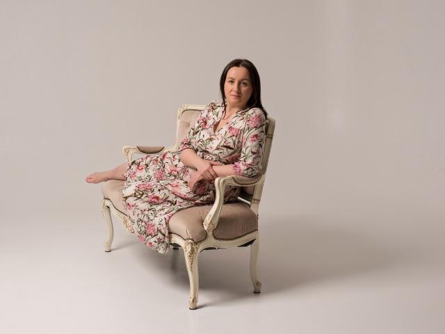 AbbyTaylorr, sitting on an ornate, beige, floral-patterned dress, with pink and green flowers, on a vintage, cream-colored, upholstered chair, in a neutral studio setting.