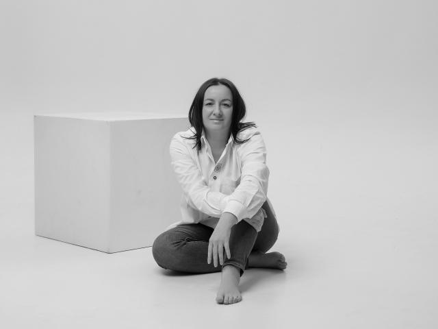 AbbyTaylorr, pieds nus, assise en tailleur, portant une chemise blanche à boutons et un pantalon foncé, souriante, à côté d'un cube blanc, photo en noir et blanc, cheveux foncés de longueur moyenne, détendue..