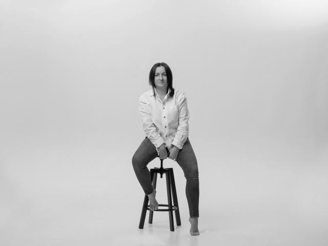 AbbyTaylorr zit op een kruk, gekleed in een overhemd en jeans, met haar billen iets gespreid, en een rustige uitdrukking, op een zwart-wit foto..