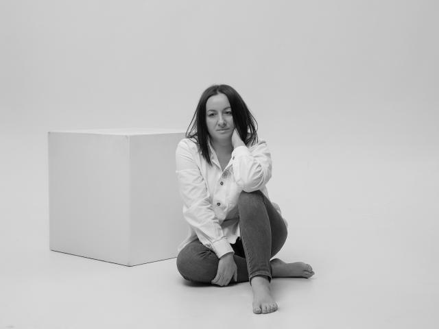 AbbyTaylorr with tits covered, wearing a button-up shirt, jeans, and bare feet, sitting next to a cube, looking down with a slight smile, in a black-and-white photo.
