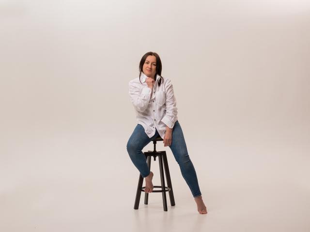 AbbyTaylorr sits barefoot on a stool, wearing a white button-up shirt and blue jeans, with her hand near her mouth, looking slightly to the side.