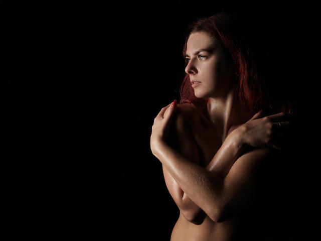 MelanyTessk a la peau brillante et humide, des cheveux roux et de petites seins, qu'elle couvre de ses mains, sur une photo sombre et mélancolique, avec une expression concentrée et pensive..