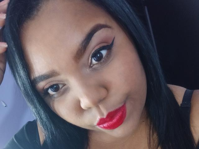 Het gezicht van SarahSmity, met donkerbruine ogen, opvallende eyeliner, rode lippenstift, zwart haar en bruine huid, kijkt iets opzij met een subtiele glimlach..
