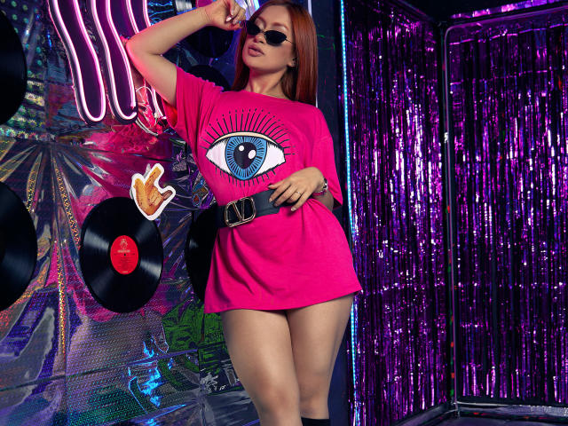 AliceSchmit with red hair, wearing a pink eye-print t-shirt, black belt, and sunglasses, standing in a neon, purple, and silver room with vinyl records and shiny curtains.
