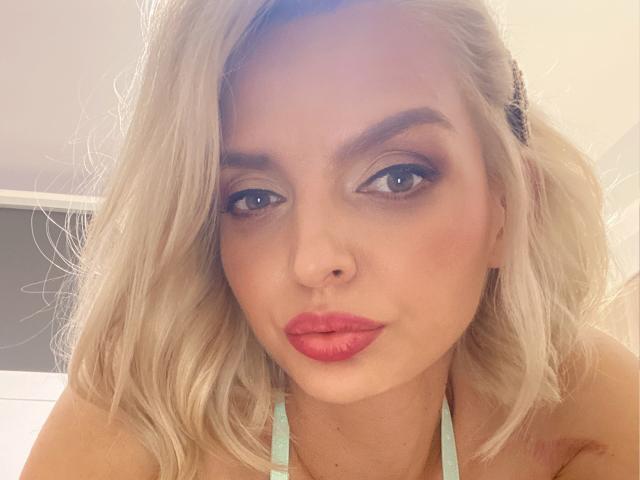 DouceAmelia has blonde hair, blue eyes, and red lipstick, with a cute nose and slightly parted pink lips. She's wearing a green halter top, looking directly at the camera.