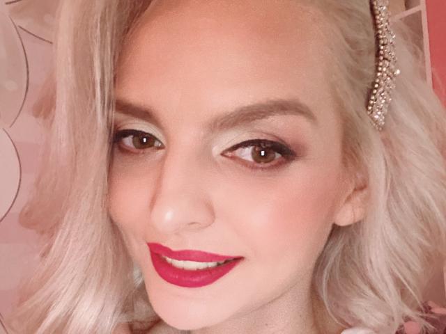 DouceAmelia's face, featuring platinum blonde hair, brown eyes, pink eyeshadow, red lipstick, and a pearl hairpiece, smiling slightly with a pinkish background.