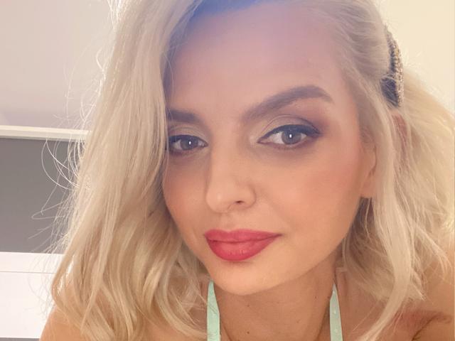 DouceAmelia has blonde hair, blue eyes, and red lipstick. Her tits are partially visible in a green halter top. She's making a slight pouty face in the selfie.