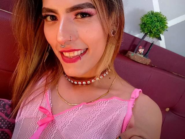 Katyling has a nose ring, lip piercing, and choker with gems, wearing pink lace, smiling with pink lipstick, and has a cute face with long brown hair.