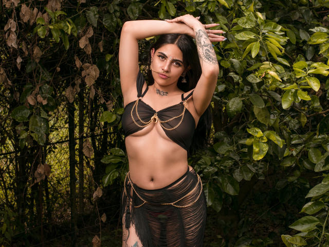 CoriPaige with dark hair, tan skin, and tattoos, wearing a black sheer bra and skirt with gold chains, arms raised, standing in front of green leafy background.