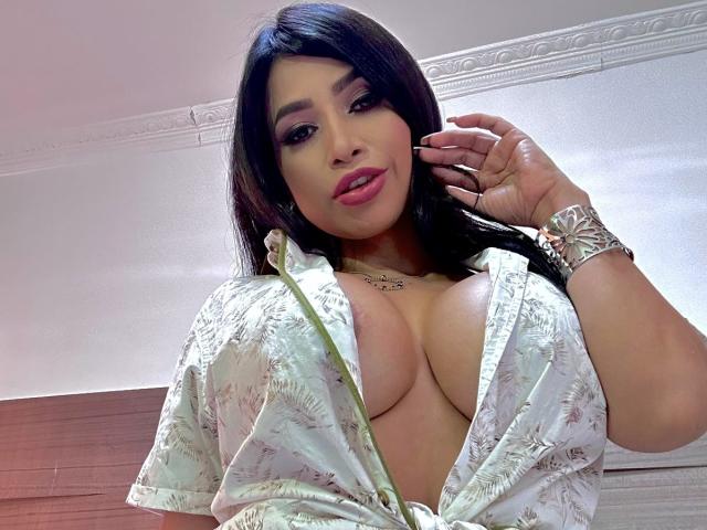 FransheskaParker with large, exposed tits in a floral, unbuttoned shirt, dark hair, pink lipstick, silver bracelet, and necklace, looking at camera with hand near head.