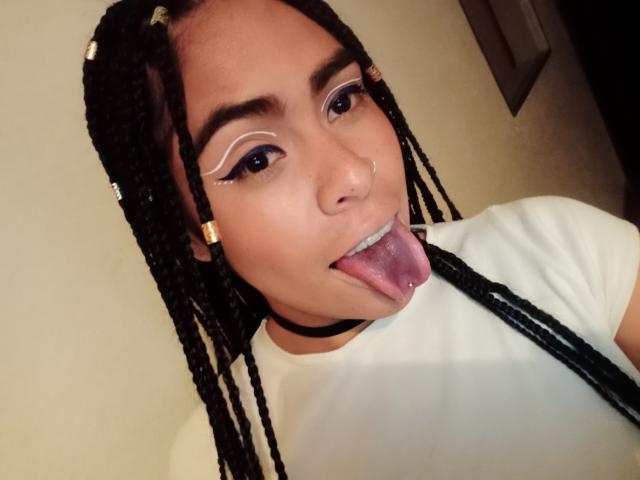 StellaBeckker has braided black hair, white winged eyeliner, and a black choker. She's sticking her tongue out, showing her pink tongue and a small nose piercing.