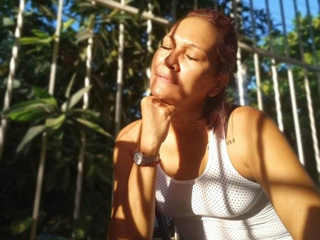 MatureJennyX with eyes closed, sunlit face, white polka-dot tank top, red hair, wristwatch, hand on chin, green leaves and white fence in background. Sunlight casts shadows on her.