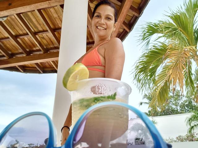 MatureJennyX with tan skin, brown hair in a bun, smiling, wearing an orange bikini, standing under a wooden roof, holding a drink with a lime slice and blue sunglasses.