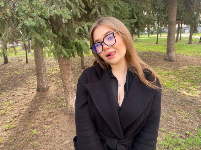 Stacy Yellig, avec des lunettes, la langue tirée, un manteau noir, décolleté, de longs cheveux blonds, debout dans une forêt, regardant la caméra, herbe verte, pins, sol légèrement sale..