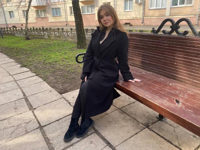 Stacy Yellig, avec ses longs cheveux bruns, est assise sur un banc en bois, vêtue d'une robe noire et de bottes noires. Ses fesses reposent sur le banc, et ses bras sont posés sur ses genoux..