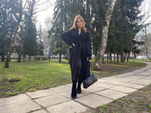 Stacy Yellig, con cabello rubio, vestida con un abrigo y pantalones negros, botas negras, y sosteniendo un bolso negro, está de pie en un camino de piedra en un parque, sonriendo con confianza..