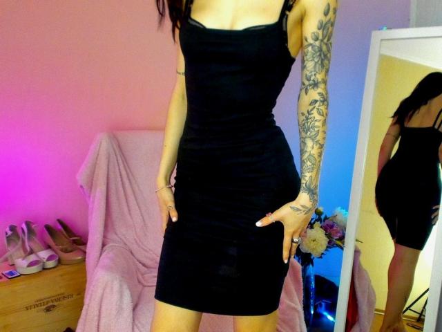 Tsukerberg in a tight black dress, showing off tattooed arm, with hands on her ass, reflected in a mirror, pink and blue lights, standing in a room.