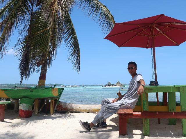 ElChicletv está relajado en un banco rojo, vistiendo pantalones grises y zapatillas negras, con un paraguas rojo y un banco verde debajo de una palmera, junto al mar azul..
