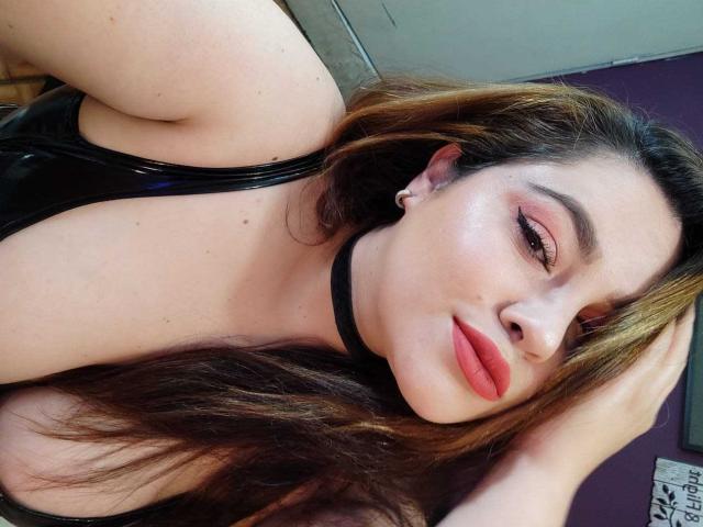LissaClaire with long brown hair, black choker, and red lipstick, wearing a black leather top, showing her boobs and smiling at the camera, hand in her hair.