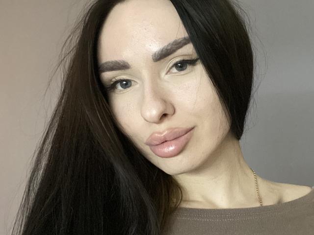 BellaMegan, con cabello largo y castaño oscuro, cejas pobladas y labios rosados y brillantes, vestida con una blusa marrón, tiene un tono de piel claro y un collar de oro discreto..