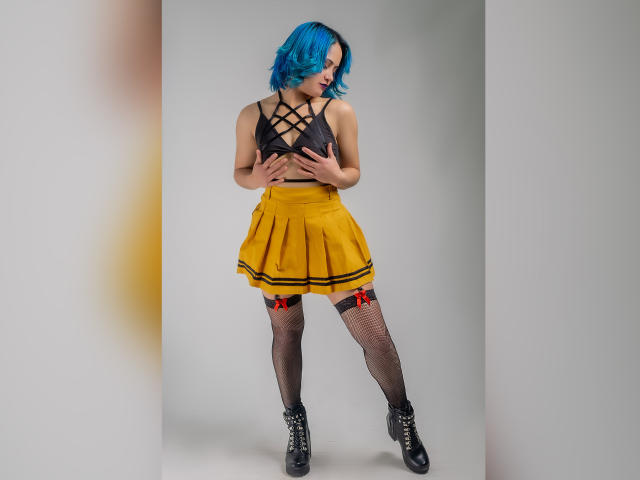 CassieJones with blue hair, black strappy top, yellow skirt, fishnet stockings with red bows, black boots. Hands on chest, looking down, standing, cute, sexy.