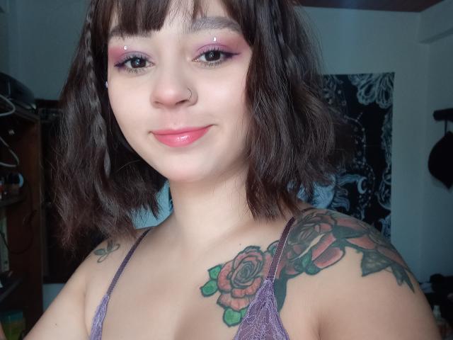 MefistoErica, con tatuaggi di rose sulle spalle, eyeliner viola, piercing al naso e capelli castani corti, sorride con le labbra rosa, indossando una camicetta viola in pizzo..