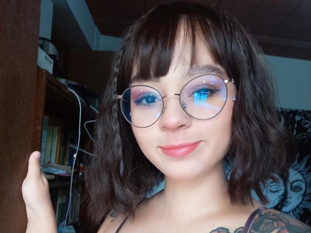 MefistoErica ha i capelli castano scuro, lunghi fino alle spalle, occhiali rotondi con occhi azzurri che riflettono la luce, un piercing al naso e un piercing al labbro. Sorride, con il rossetto rosa, mostrando un tatuaggio sul braccio..