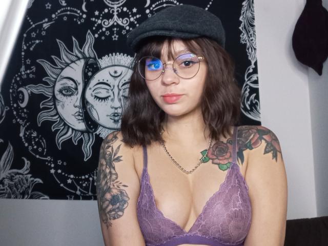 MefistoErica con seni di media grandezza, indossa un reggiseno di pizzo viola, un berretto nero, occhiali, tatuaggi sulle braccia e un tatuaggio di una rosa sul petto, una collana a catena, capelli castani ondulati, guarda verso la fotocamera..