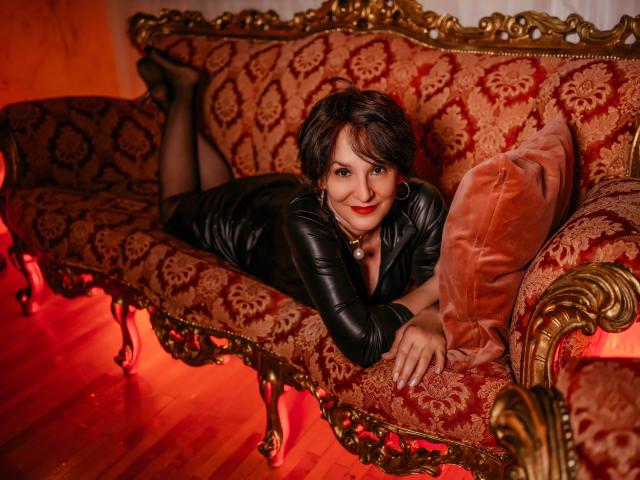 AlbertaAllen, lying on an ornate red and gold patterned couch, wearing a black leather dress, black stockings, and red lipstick, with a cute smile and short dark hair.