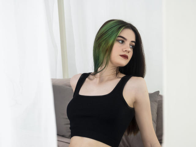 Nicole, mit grünen und schwarzen Haaren, trägt ein schwarzes Tanktop, das ihre kleinen Brüste und ihre helle Haut zeigt. Sie sitzt auf einem grauen Sofa und blickt zur Seite..