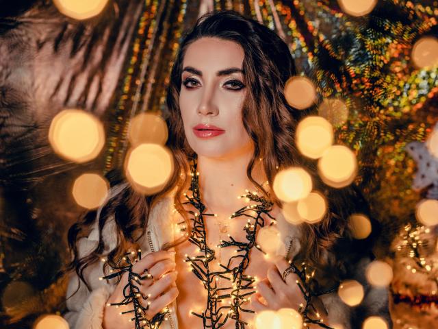 StassiLyanne, con cabello castaño ondulado, ojos brillantes y labios rojos, sosteniendo luces de Navidad frente a un fondo borroso de luces cálidas, vistiendo una blusa blanca esponjosa..