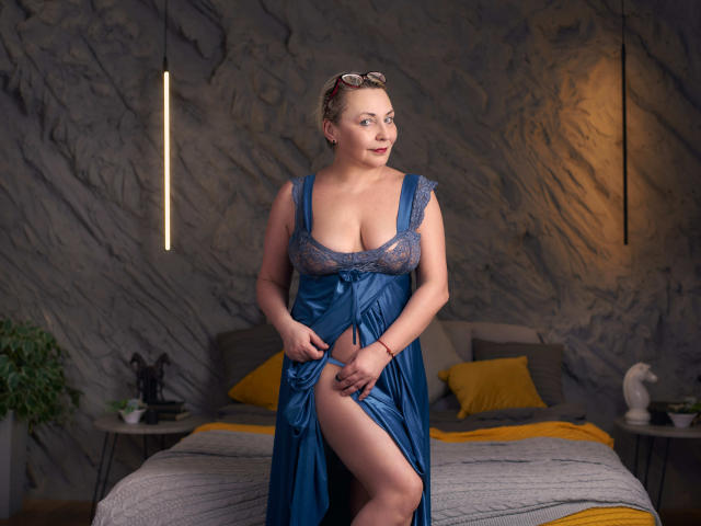 LadyIren mit großen Brüsten, trägt ein blaues Spitzen- und Satinnachthemd, zieht ihren Rock hoch, um ihren Oberschenkel zu zeigen, und steht in einem Schlafzimmer mit gelben Kissen..