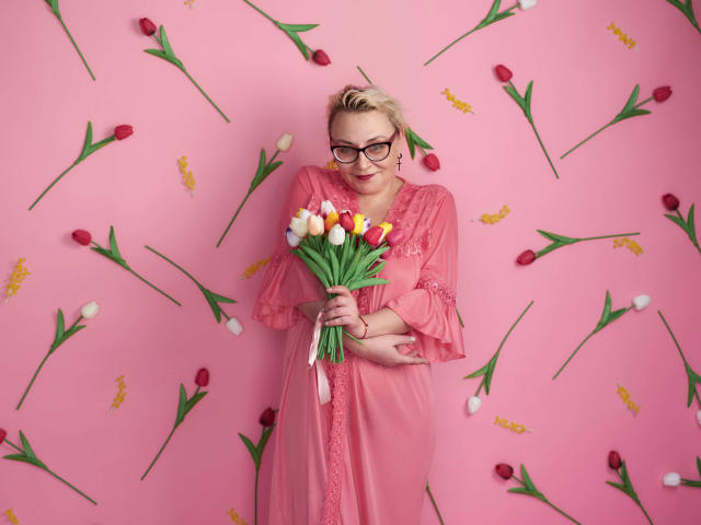 LadyIren mit blonden Haaren, Brille und einem pinken Kleid, hält einen Strauß aus roten, weißen und gelben Tulpen und steht vor einer rosa Wand, auf der Tulpen verstreut liegen..