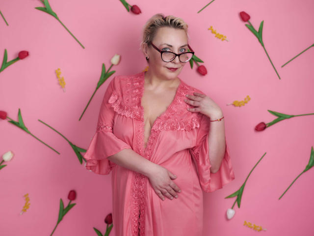 LadyIren mit blonden Haaren, Brille und einem rosa Spitzenkleid, die ihre Brüste berührt, steht vor einer rosa Wand, auf der sich rote und gelbe Tulpen befinden..