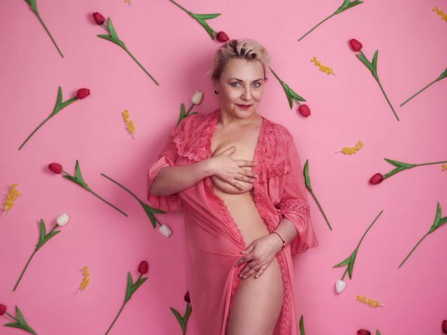 LadyIren mit kurzen, blonden Haaren, die einen rosa Spitzenmantel trägt, wobei sie ihre Brüste mit einer Hand bedeckt und die andere Hand auf ihrer rosa Vulva ruht, umgeben von roten und weißen Tulpen..