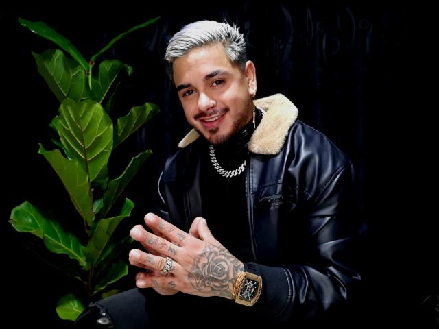 VisiI con capelli grigi, giacca di pelle nera e colletto di pelliccia color cuoio, mano tatuata, anello e orologio, sorridente, pianta verde a sinistra, sfondo nero..