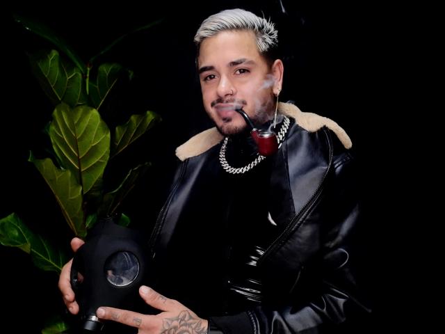 VisiI avec des cheveux courts et teints en blanc, une veste en cuir noir, un collier en chaîne, tenant une pipe, en train de fumer, une plante verte sur la gauche, un tatouage sur la main droite, fond noir..