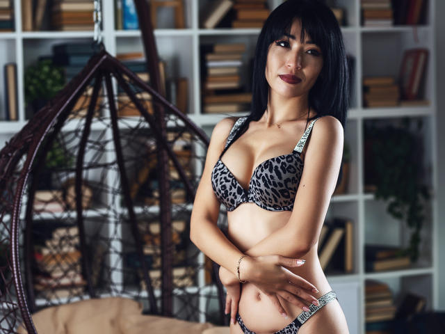 LilaBabeX with black bob, wearing leopard-print bra and panties, standing in room, arms crossed, hands on belly, brown eyes, red lipstick, medium tits, cute, confident, sexy.