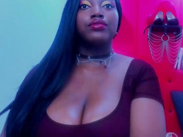 RubySwitch com pele marrom escura, cabelo preto longo, sombra amarela, batom vermelho, seios grandes, decote, blusa marrom com um ombro descoberto, colar de corrente e expressão confiante..