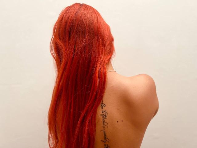 LisaHarvie, met lang rood haar, ongetooid, met een tatoeage zichtbaar aan de rechterkant, kleine zwarte vlekjes in de buurt van haar schouderblad, op een effen witte achtergrond..