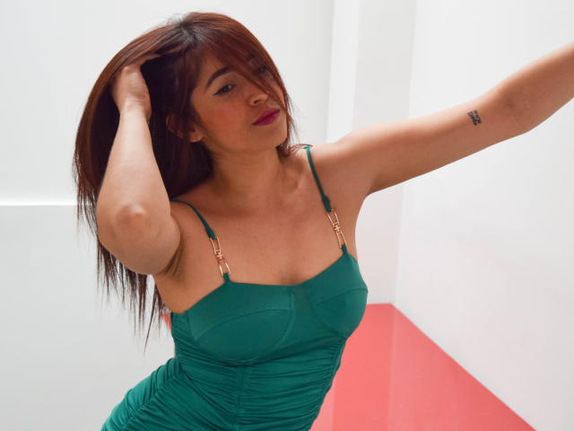 NicoColeman with brown hair, green dress, hands in hair, small tits, red lipstick, tattoo on right arm, looking at camera, white and red wall in background.