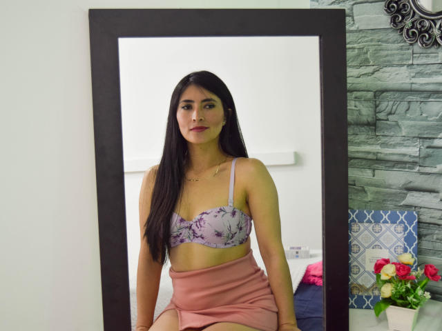 JainaRogers with long black hair, wearing a purple floral bra, showing her tits, and a pink skirt, sitting in front of a mirror, looking serious.