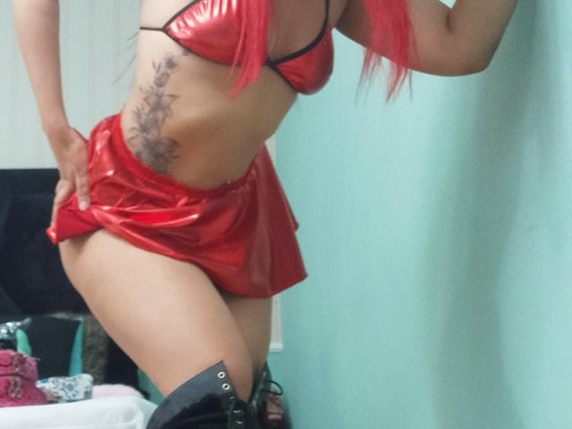 TarahDustin with red shiny bikini top and skirt, black boots, and red hair, showing a tattoo on her side, her ass, and her small tits.