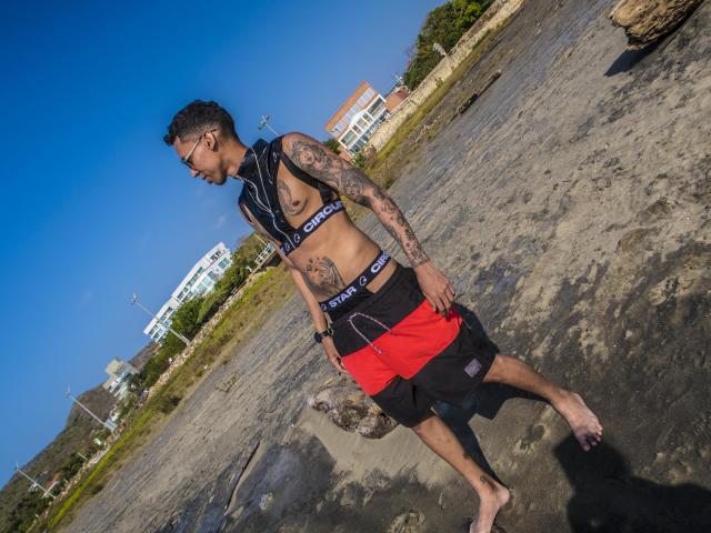 ElChicletv, sin camisa, con tatuajes en los brazos y el pecho, pantalones cortos negros y rojos, de pie en una playa rocosa, descalzo, mirando hacia abajo, cielo azul claro, de día, cabello corto y oscuro, gafas..