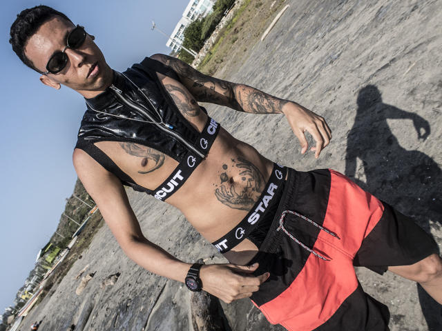 ElChicletv est un homme tatoué, avec des cheveux courts et foncés, portant des lunettes de soleil, un haut noir brillant sans manches et un short rouge et noir, debout sur une plage rocheuse avec une montre noire au poignet..