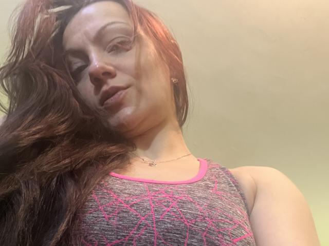 AamyBeatrice with long brown hair, wearing a pink and gray sports top, showing her tits, slight smile, necklace, and light skin, in a low-angle selfie.