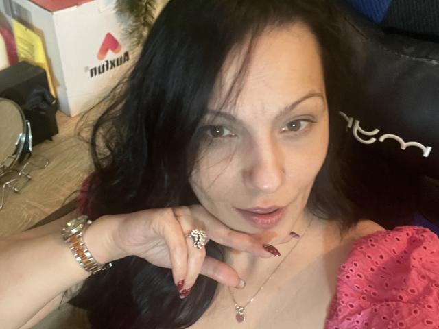 AamyBeatrice met lang zwart haar, groene ogen en rode nagels, die een roze top draagt, een gouden armband en een ring, en haar nek aanraakt, terwijl ze naar de camera kijkt..