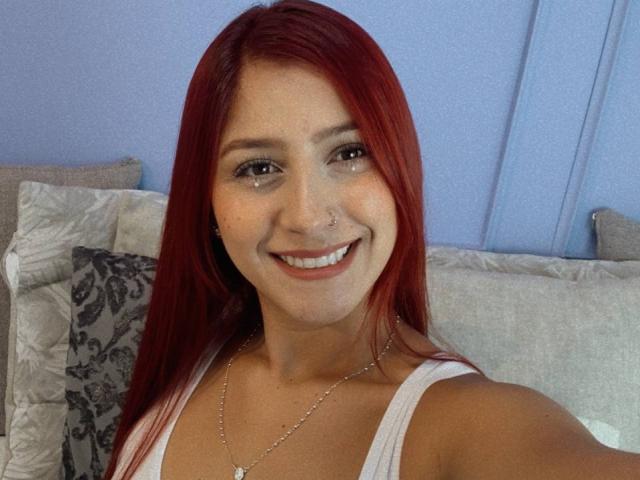 LisaHarvie, com cabelo ruivo, sorrindo, com piercing no nariz, vestindo uma camiseta branca, com um colar, segurando um celular, com os seios visíveis, fofa, pele clara, olhos castanhos, sentada na cama com travesseiros..